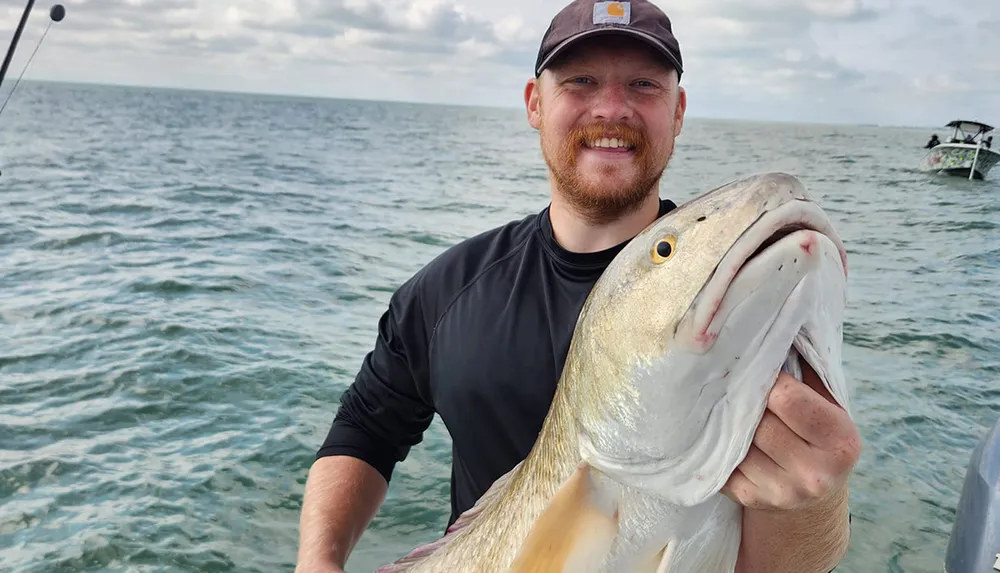 A smiling person proudly holds up a large fish they caught with the sea in the background