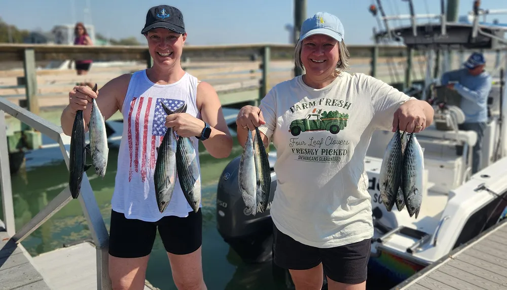 Two women are smiling and holding up fish on a sunny dock showcasing their catch from a fishing trip