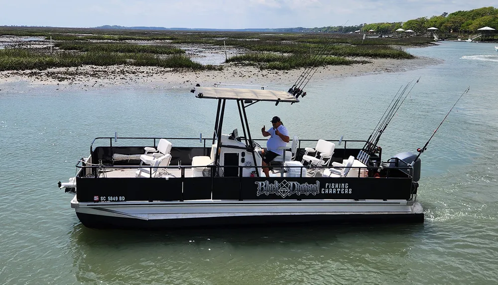 A person is standing on a fishing charter boat equipped with rods navigating through shallow waters near a marshy area