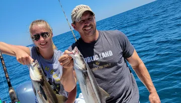 A man and a woman are smiling and posing with two freshly caught fish on a boat with the ocean in the background.