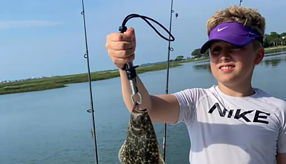 A young person wearing sunglasses and a baseball cap holds up a fish they have caught with a tranquil waterway and greenery in the background