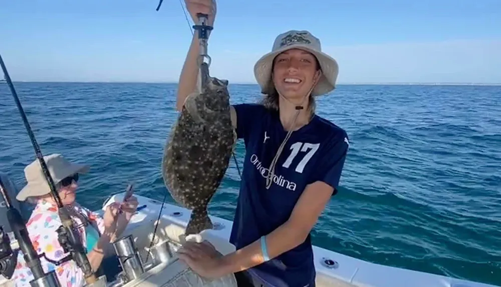 A joyful person is holding up a fish they caught on a sunny day at sea while another person seated on the boat looks on