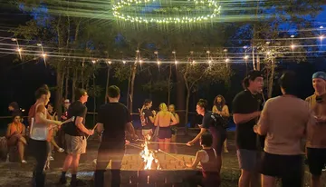 People are gathered around a campfire at twilight, enjoying conversation and roasting marshmallows under a canopy of string lights.