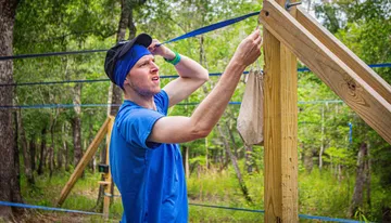 A person in a blue shirt and headband appears to be engaged in an outdoor physical activity or construction task, wiping sweat from their forehead near a wooden structure with hanging ropes or straps.
