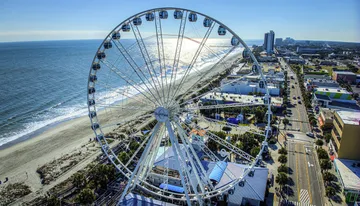 A large Ferris wheel dominates the foreground with a view of a coastal cityscape and beach in bright daylight.