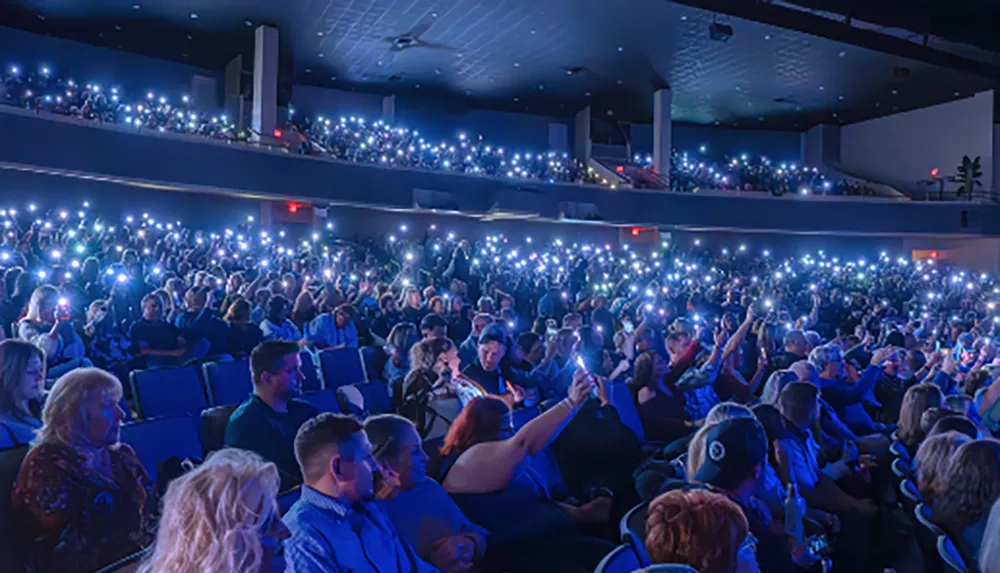 A large audience in a theater is holding up lit cell phones creating a sea of lights