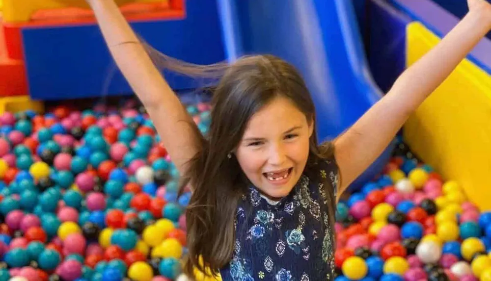 A child joyfully plays in a colorful ball pit raising their arms in excitement
