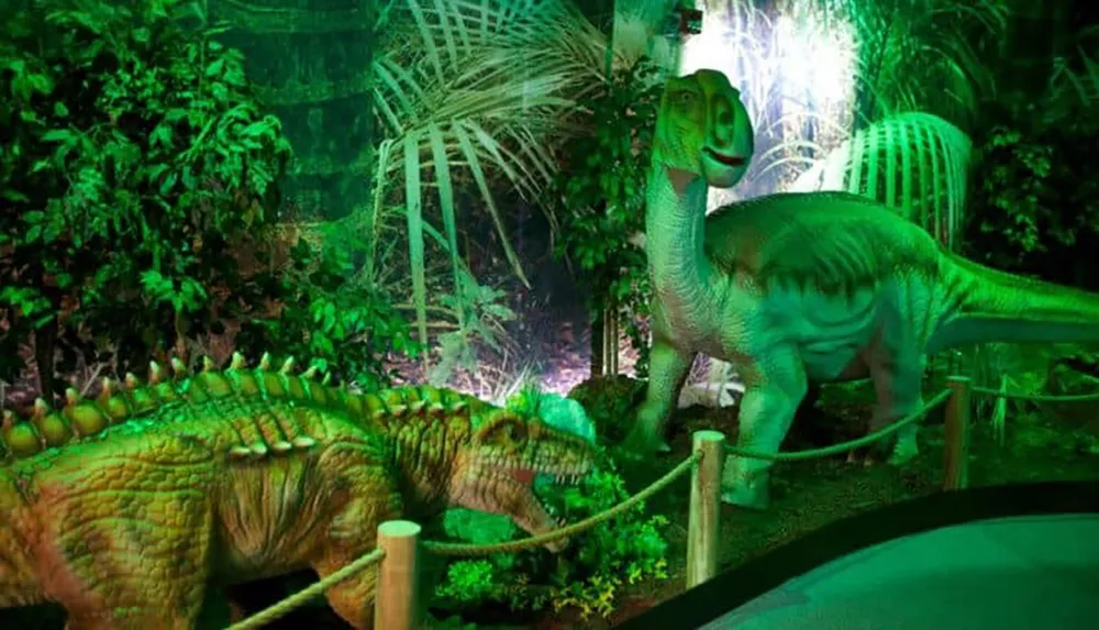 The image depicts two dinosaur models in a lush green forest-like setting with artificial lighting