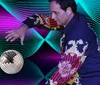 A person in a colorful ornate jacket poses dramatically next to a disco ball with a vibrant abstract background