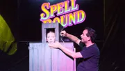 A magician performs a trick involving a woman in a box, with the word 