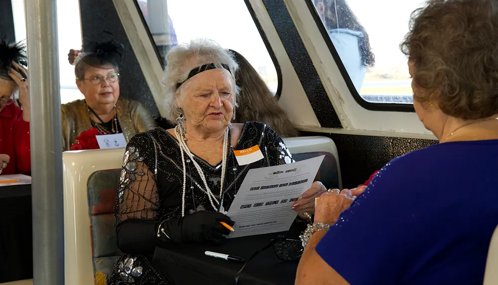 An older woman dressed in vintage-style clothing is studying a paper while seated at a table with another person on a boat