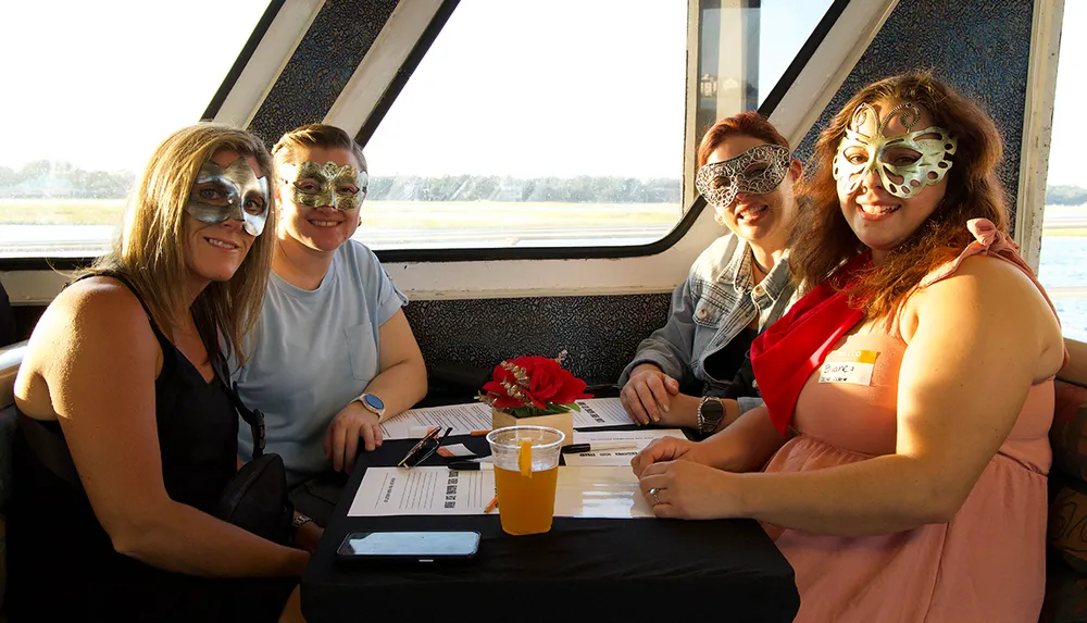 Four people wearing decorative masks are sitting at a table on a boat with drinks and a view of the water in the background
