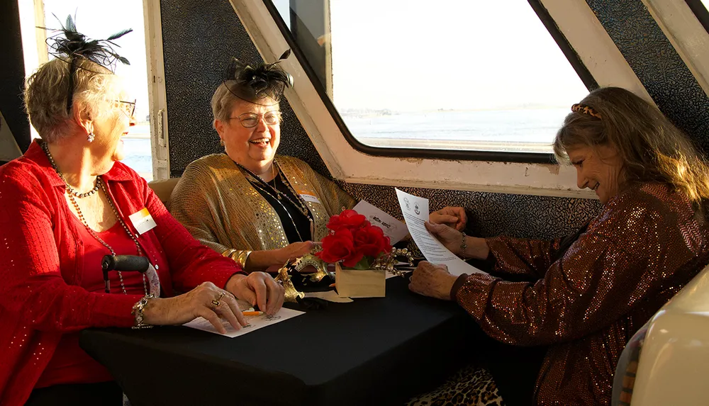 Three women dressed in sparkly clothes with fascinators are joyfully chatting and reading a paper at a table by a window