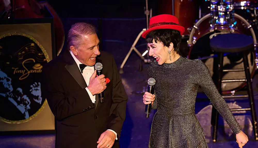A man in a tuxedo and a woman in a sparkly dress with a red hat are singing together on stage