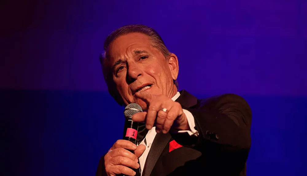 A man in a tuxedo is holding a microphone and gesturing with his hand while on stage