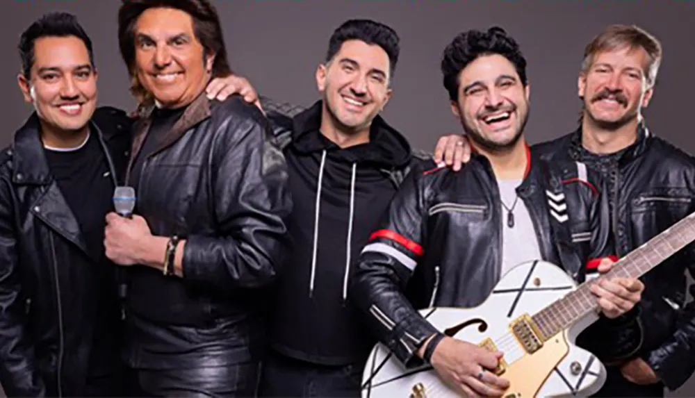 Five men are posing together with one holding a guitar and another holding a microphone all dressed in casual black and leather attire