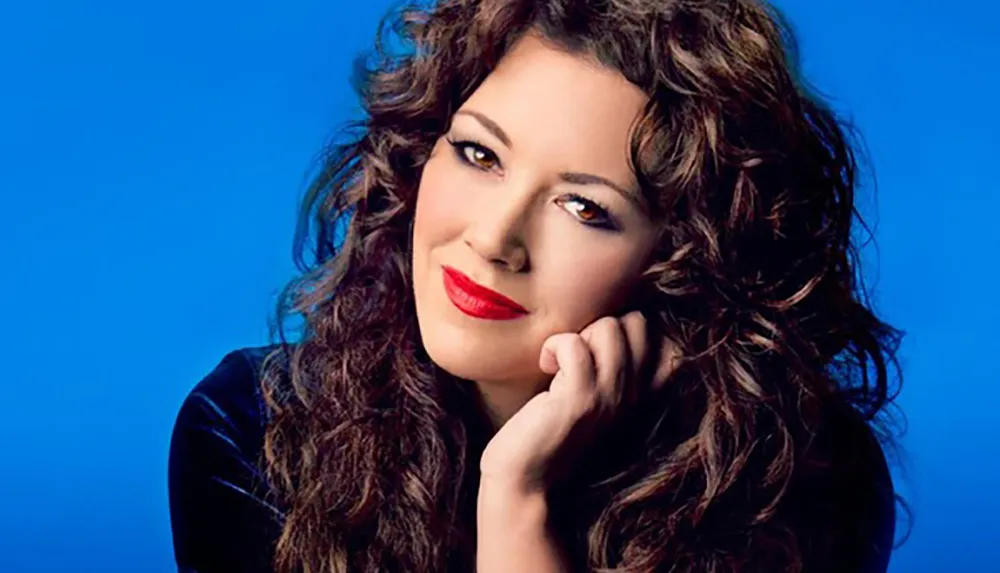 A woman with curly hair and red lipstick is posing against a blue background resting her chin on her hand