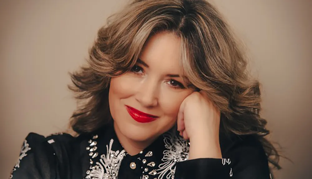 A person with wavy hair and red lipstick is posing with their hand on their face wearing a black garment with intricate white embroidery