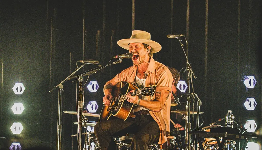 A musician passionately sings and plays guitar on stage wearing a wide-brimmed hat surrounded by musical equipment and stage lights