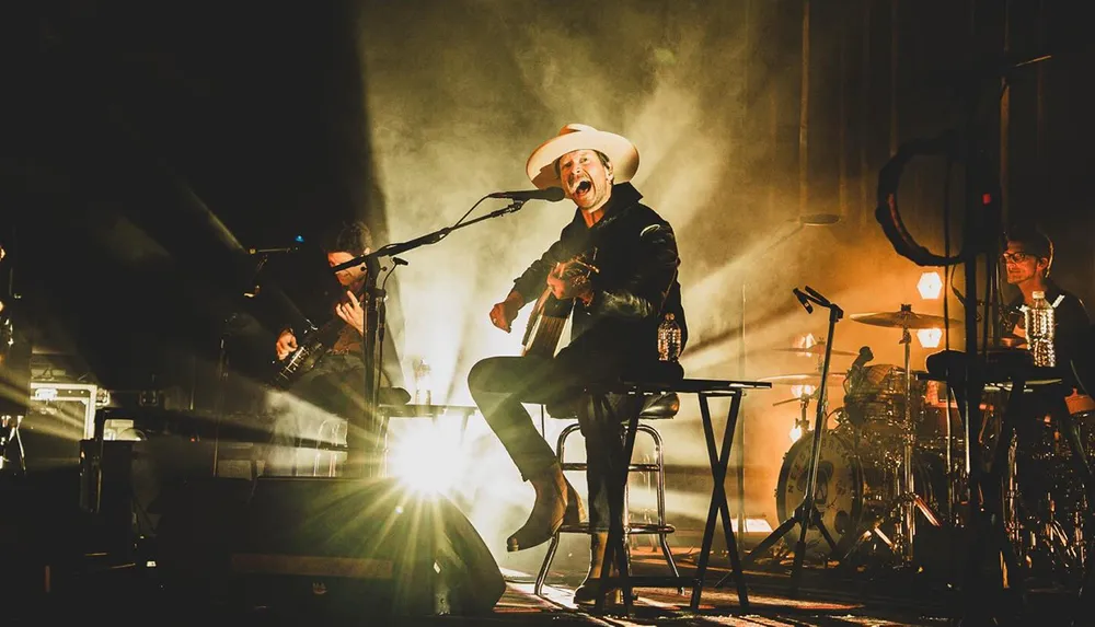 A musician wearing a hat passionately performs on stage surrounded by band members and illuminated by dramatic backlighting