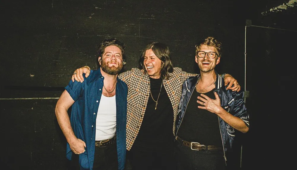 Three people are happily posing together with their arms around each other in front of a dark background