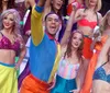 A group of performers in vibrant colorful disco outfits are enthusiastically singing on a stage with a bright multicolored background