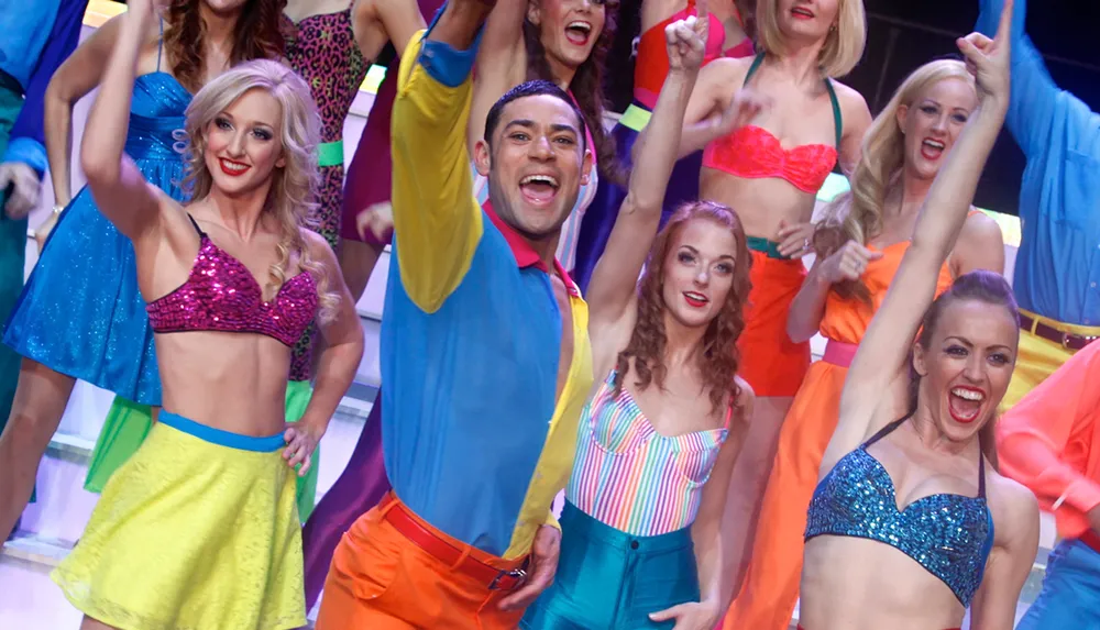 A group of performers in colorful vibrant costumes is energetically posing with their arms raised and smiling on stage