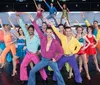 A group of performers in vibrant colorful disco outfits are enthusiastically singing on a stage with a bright multicolored background