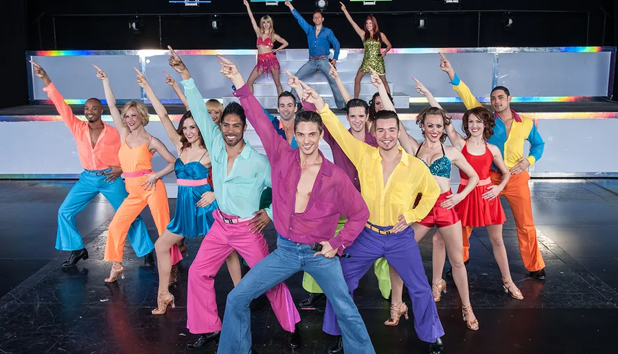 A large group of dancers in vibrant, colorful costumes strike energetic poses on stage.