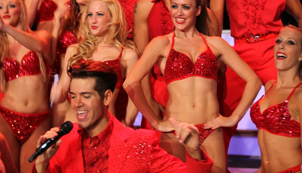 A group of performers in red sequin costumes are energetically singing and dancing on stage