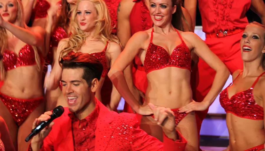 A group of performers in red sequin costumes are energetically singing and dancing on stage.