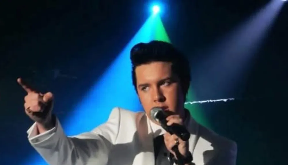A person in a white suit is performing on stage holding a microphone and gesturing with their hand while colorful stage lights shine in the background