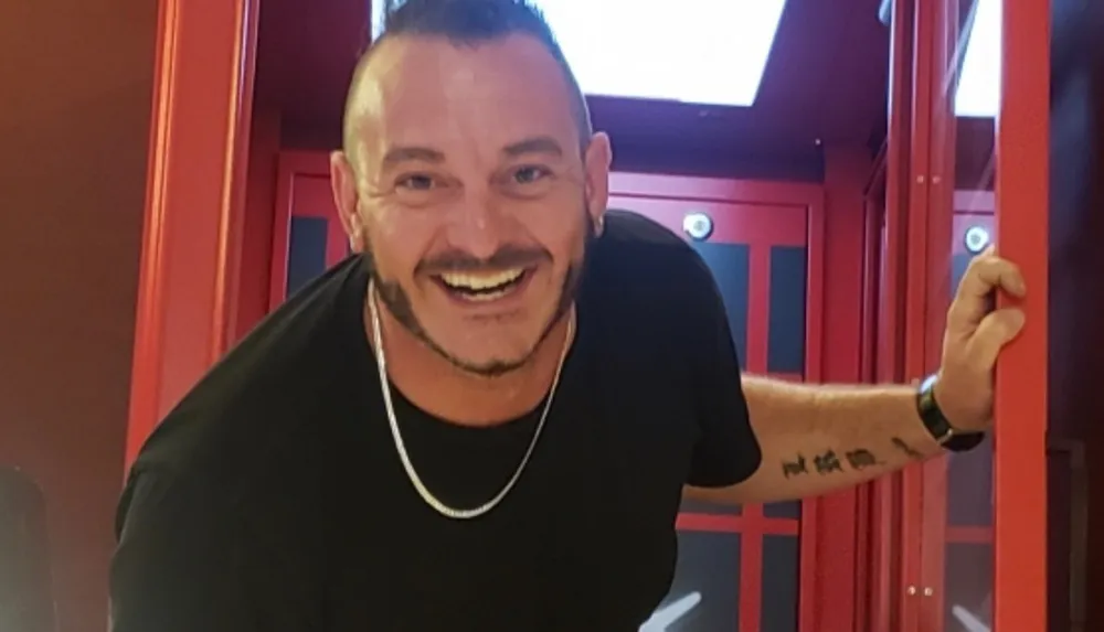 A smiling man with a beard is standing in a brightly lit room holding onto a red doorframe