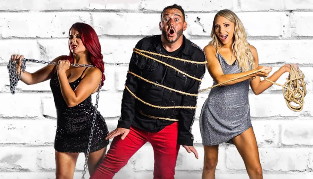 Three people are standing in front of a white brick wall with the man in the middle tied up in rope and women on either side holding chains and rope looking surprised and amused