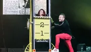 A woman looks surprised inside a box marked with numbers, while a smiling man interacts with the setup during a likely magic performance.