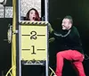 A woman looks surprised inside a box marked with numbers while a smiling man interacts with the setup during a likely magic performance