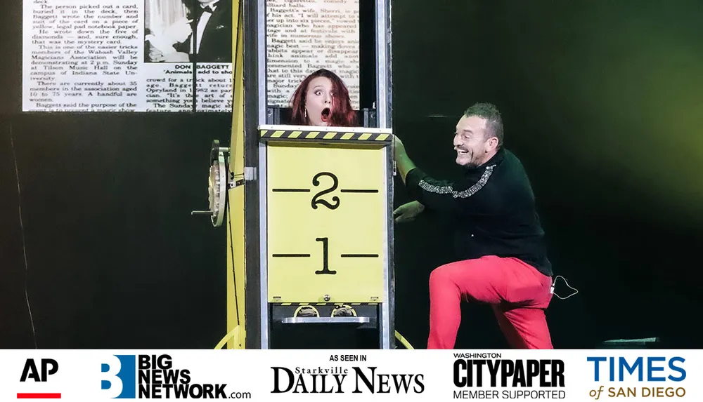 A woman looks surprised inside a box marked with numbers while a smiling man interacts with the setup during a likely magic performance