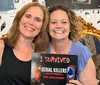 Two smiling women are sitting together holding a sign that reads I Survived Serial Killers The Experience True Crime Museum