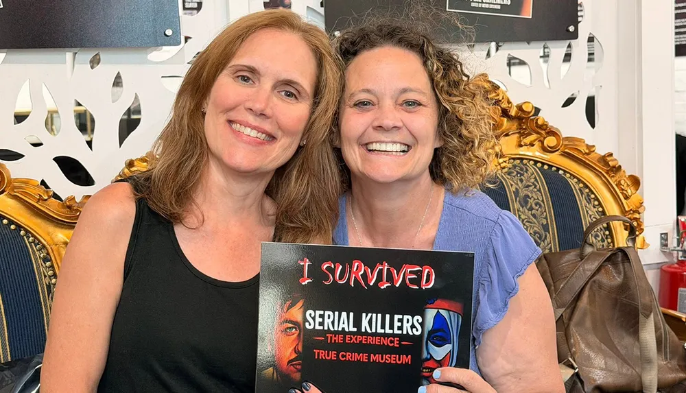 Two smiling women are sitting together holding a sign that reads I Survived Serial Killers The Experience True Crime Museum