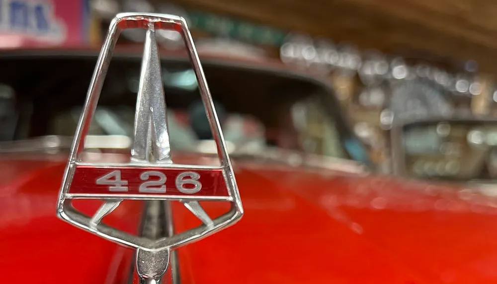 A close-up of a cars hood ornament displays the number 426 on a red surface with a blurred background