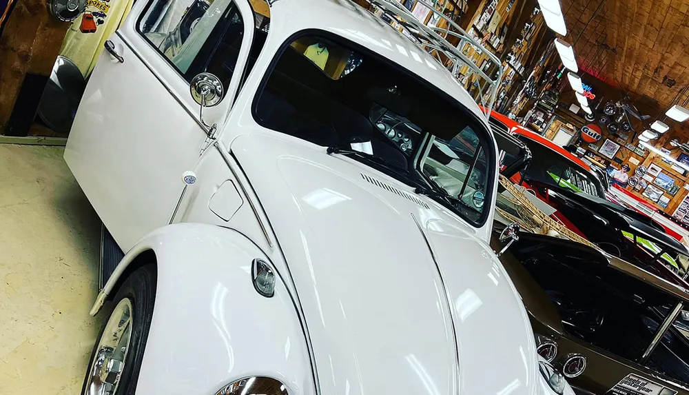 A white vintage car is parked in a garage filled with various classic cars and automotive memorabilia
