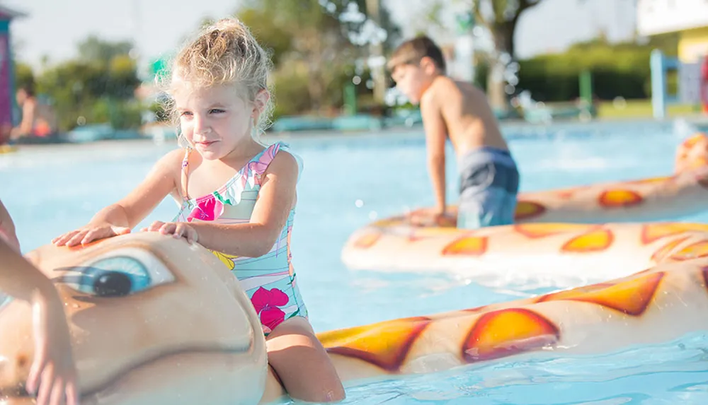 A young girl in a swimsuit is playing on a turtle-shaped float in a pool with another child nearby