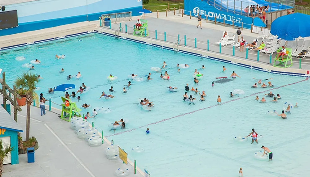 The image shows a busy outdoor wave pool where people are enjoying the water some floating on inflatable rings with a Flowrider surf simulator visible in the background