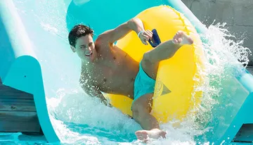 A person is joyfully sliding down a water slide on a yellow inflatable ring.