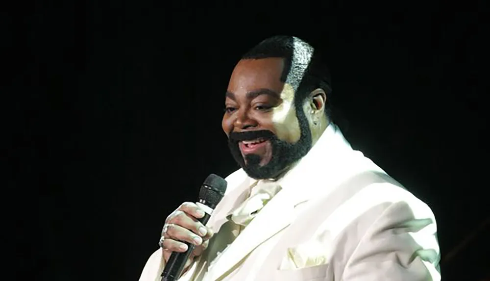 A man in a light-colored suit is holding a microphone and smiling on stage