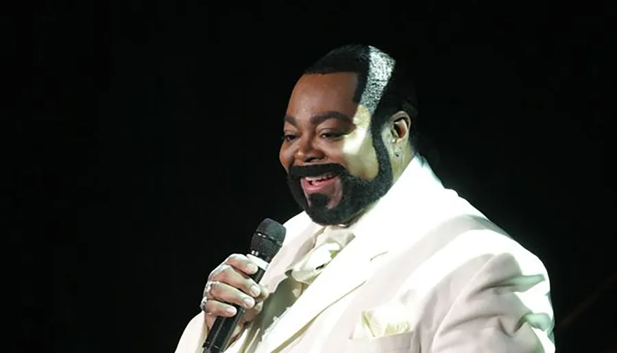 A man in a light-colored suit is holding a microphone and smiling on stage.