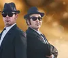 Two men dressed in black suits hats and sunglasses pose back-to-back with a blurred gold background