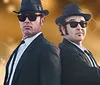 Two men dressed in black suits hats and sunglasses pose back-to-back with a blurred gold background