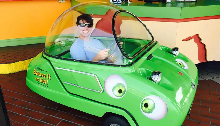 A person is smiling while sitting in a whimsical green car with large eyes, which is a part of an exhibit at Ripley's Believe It or Not! Museum.
