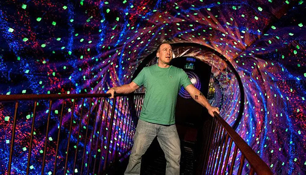 A person stands on a bridge inside a dazzling tunnel-like space with walls covered in colorful luminous patterns